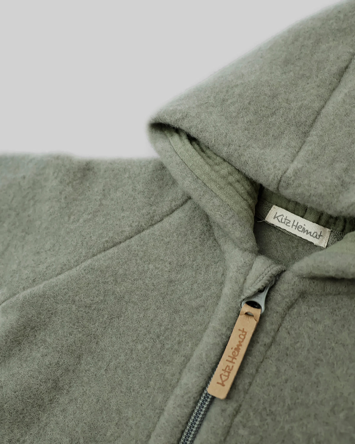 Nahaufnahme der Kitz Heimat Wollfleece-Jacke Frost Green/Green mit flauschiger Wollstruktur, Kapuzenfutter und Reißverschluss mit Markenanhänger.