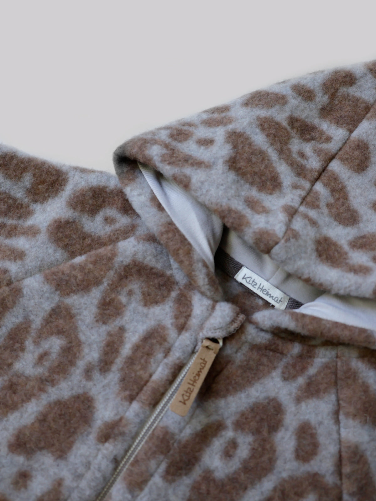 Nahaufnahme der Kitz Heimat Wollfleece-Jacke Leo Beige mit flauschigem Leopardenmuster, hellem Kapuzenfutter und Reißverschluss mit Markenanhänger.