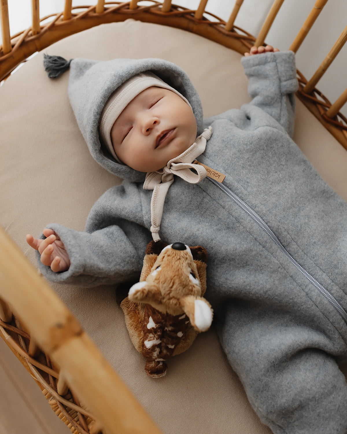 Schlafendes Baby im Kitz Heimat Wollfleece-Overall Sky Grey/Grey mit Kapuze, Quaste und Reißverschluss, liegend in einem geflochtenen Babykorb neben einem Kuscheltier.