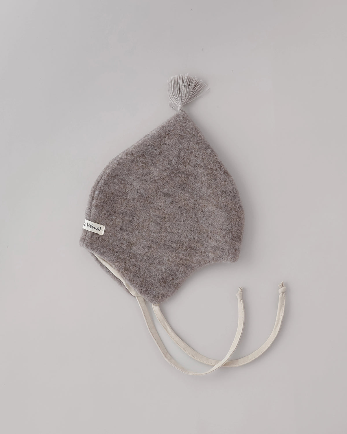 Kitz Heimat Wollfleece-Babymütze Greige/Nude mit spitzer Form, heller Quaste, cremefarbenem Futter und Bindebändern auf neutralem Hintergrund.