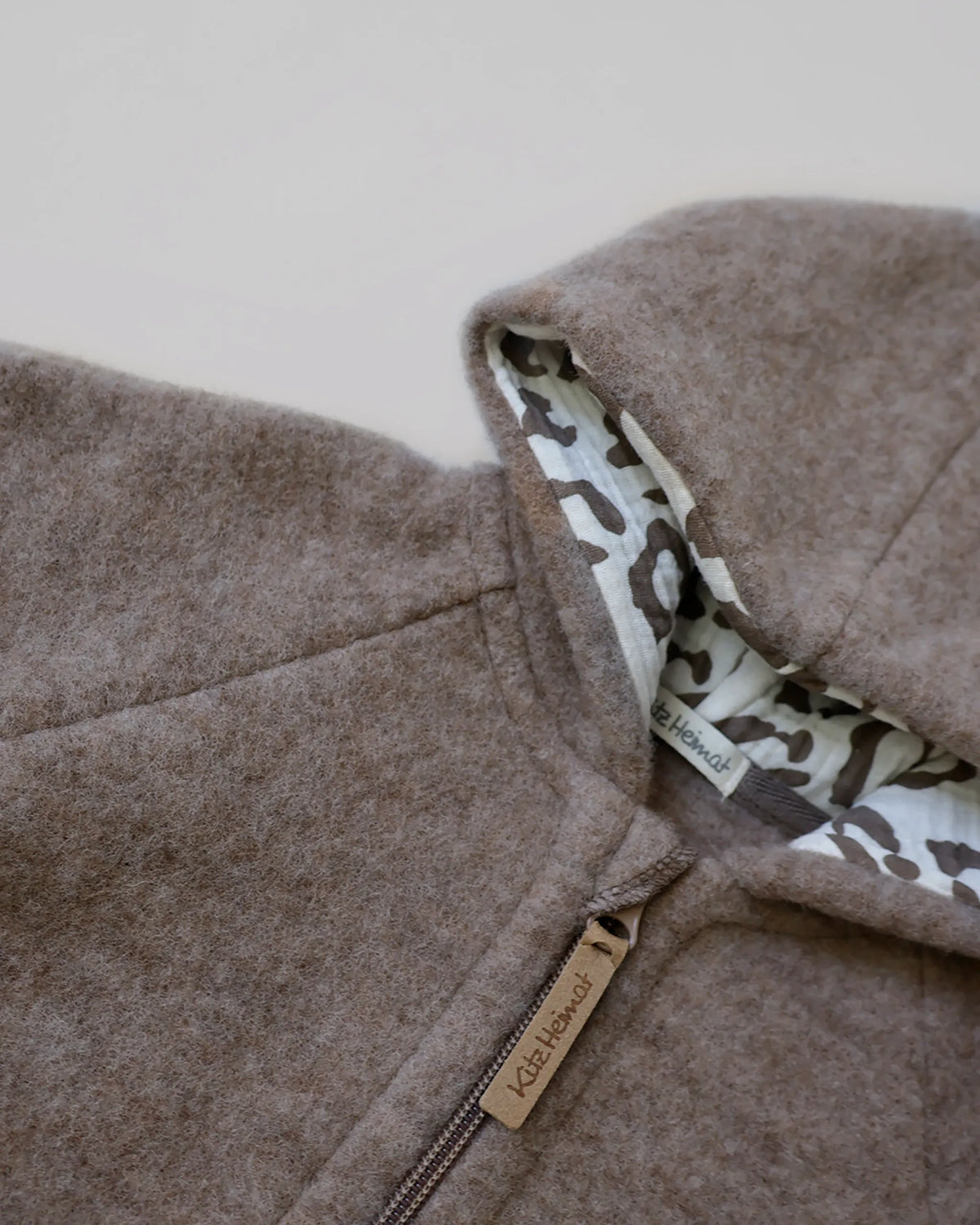 Nahaufnahme der Kitz Heimat Wollfleece-Jacke Greige/Leo mit flauschiger Wollstruktur, Kapuzenfutter im Leopardenmuster und Reißverschluss mit Markenanhänger.