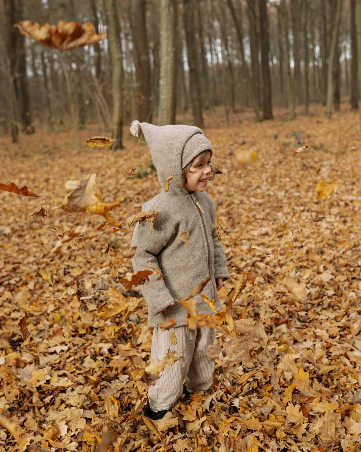 Kleinkind in der Kitz Heimat Wollfleece-Jacke Greige/Nude mit spitzer Kapuze und Reißverschluss steht zwischen fallenden Herbstblättern im Wald.