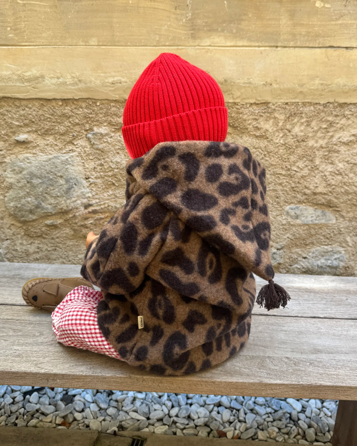 Kleinkind in der Kitz Heimat Wollfleece-Jacke Leo Coffee mit Leopardenmuster, spitzer Kapuze und dunkler Quaste, von hinten auf einer Holzbank sitzend.
