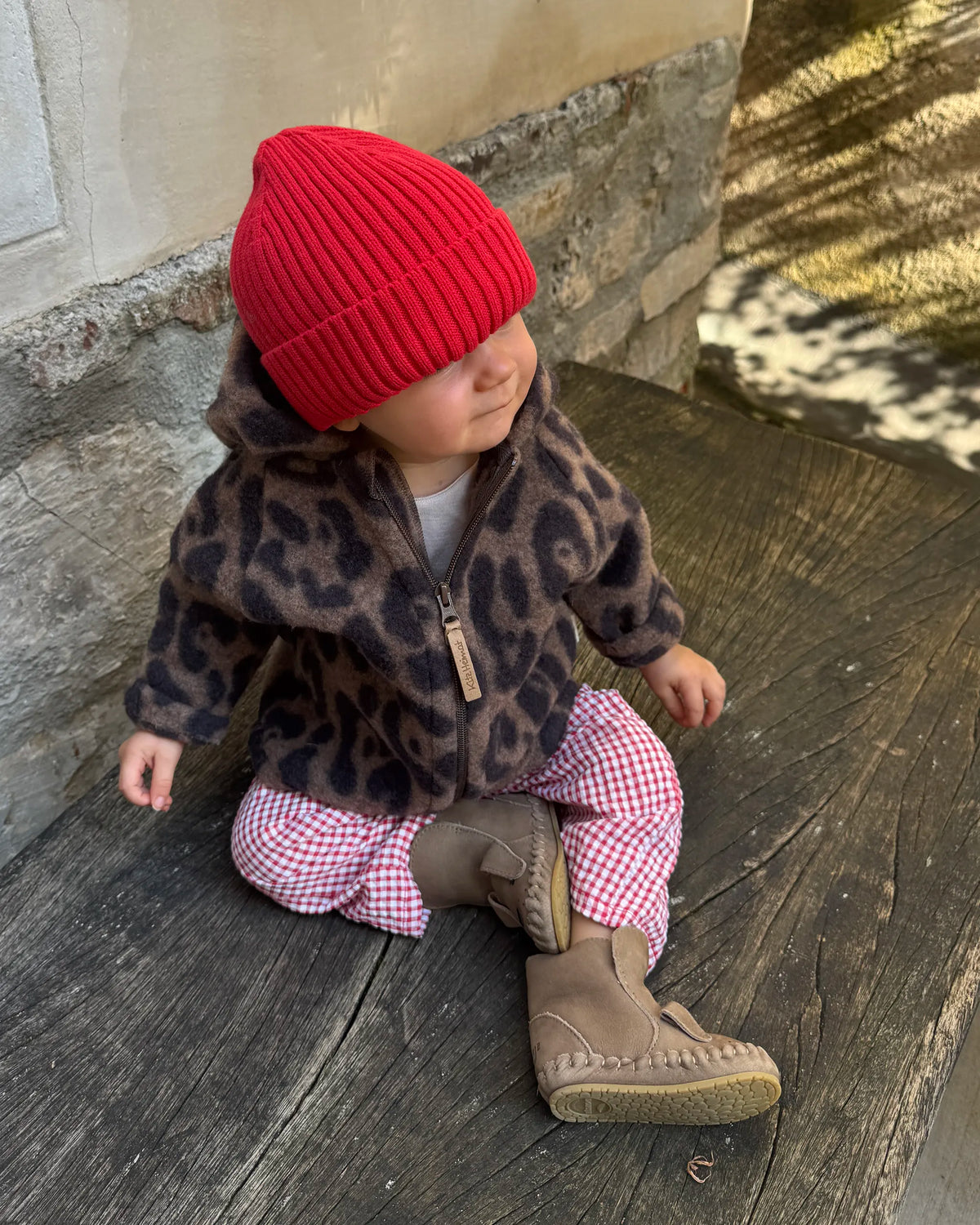 Kleinkind in der Kitz Heimat Wollfleece-Jacke Leo Coffee mit braun-schwarzem Leopardenmuster und Reißverschluss sitzt mit roter Strickmütze auf einer Holzbank.