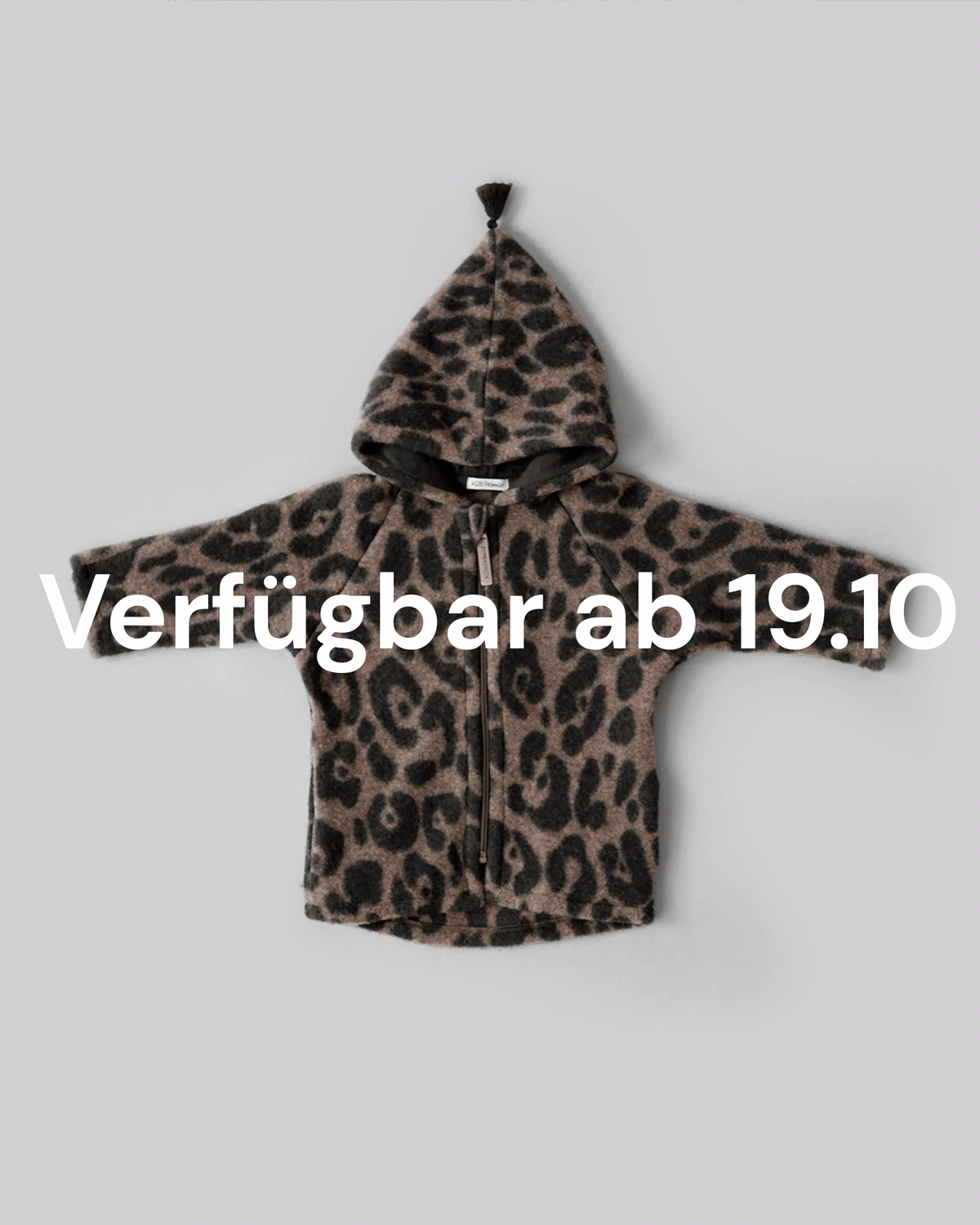 Kitz Heimat Wollfleece-Jacke Leo Coffee mit Leopardenmuster und Kapuze in Vorderansicht. Weißer Texthinweis: „Verfügbar ab 19.10“.