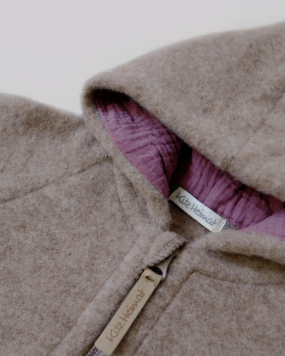 Nahaufnahme des Kitz Heimat Wollfleece-Overalls Greige/Light Plum mit flauschiger Wollstruktur, pflaumenfarbenem Kapuzenfutter und Reißverschluss mit Markenanhänger.