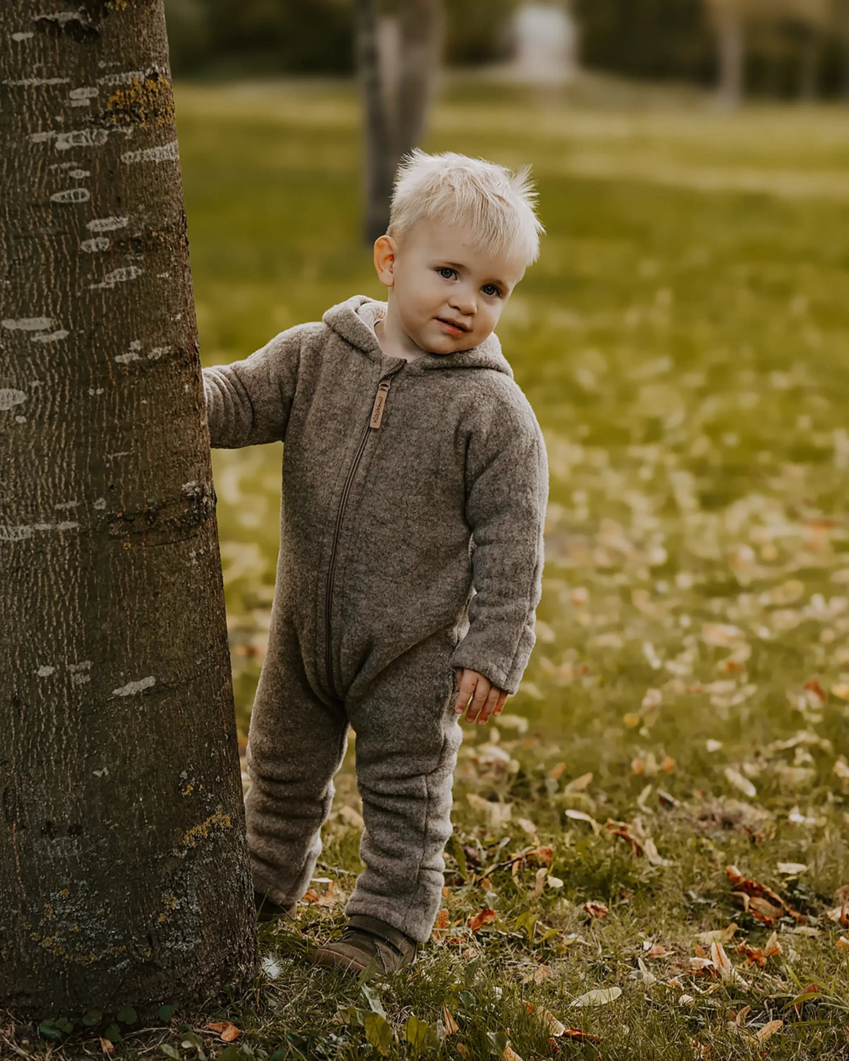 Kleinkind trägt einen Kitz Heimat Wollfleece-Overall in Greige/Nude mit Kapuze und Reißverschluss und steht neben einem Baum.