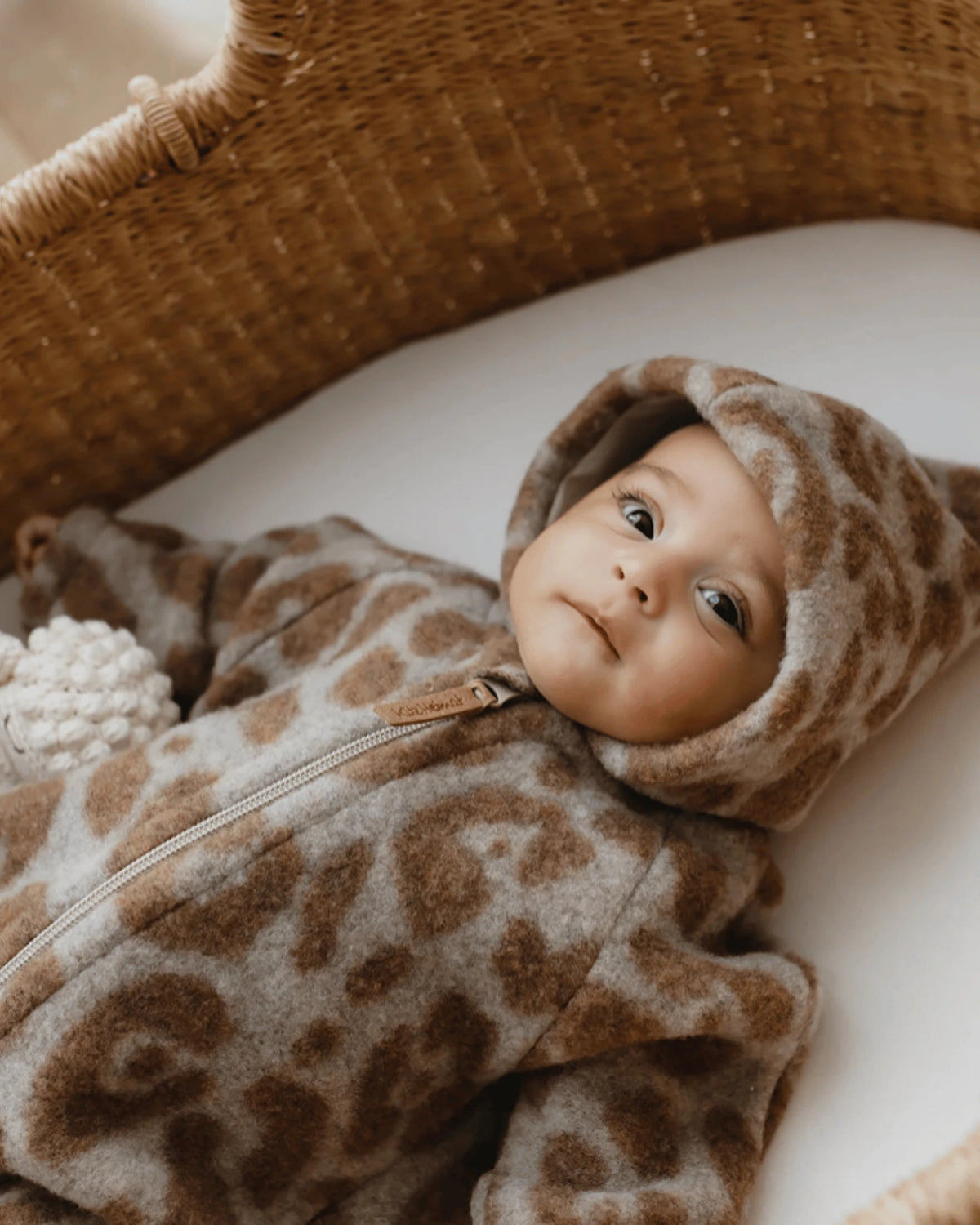 Nahaufnahme eines Babys im Kitz Heimat Wollfleece-Overall Leo Beige/Nude mit braunem Leopardenmuster und Kapuze, liegend im Babykorb.