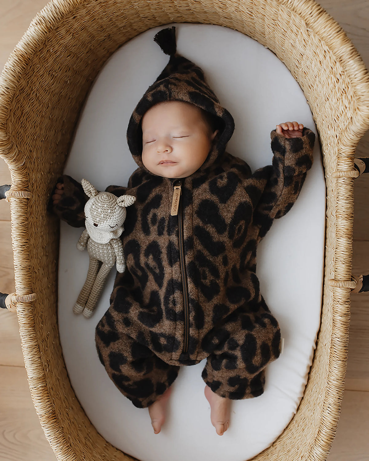 Schlafendes Baby im Kitz Heimat Wollfleece-Overall Leo Coffee mit Leopardenmuster, Kapuze und Reißverschluss in einem geflochtenen Babykorb.