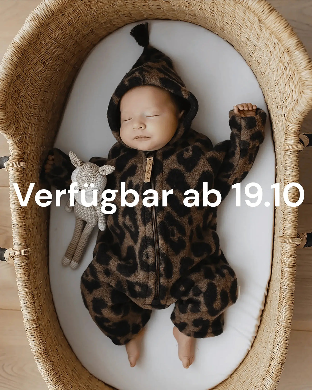 Schlafendes Baby im Kitz Heimat Wollfleece-Overall Leo Coffee/Nude mit Leopardenmuster im Babykorb. Texteinblendung: „Verfügbar ab 19.10“.