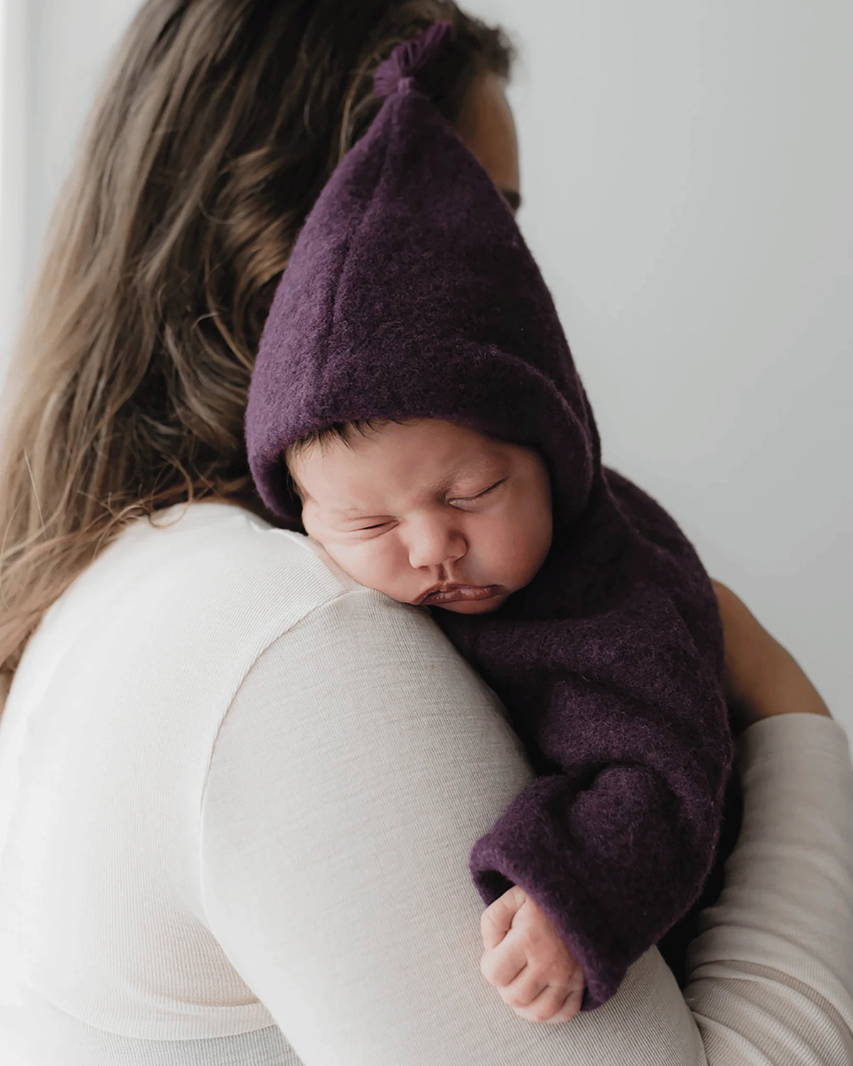 Schlafendes Baby im Kitz Heimat Wollfleece-Overall Plum/Light Plum aus Bio-Merinowolle mit spitzer Kapuze und Quaste, auf dem Arm einer Frau.