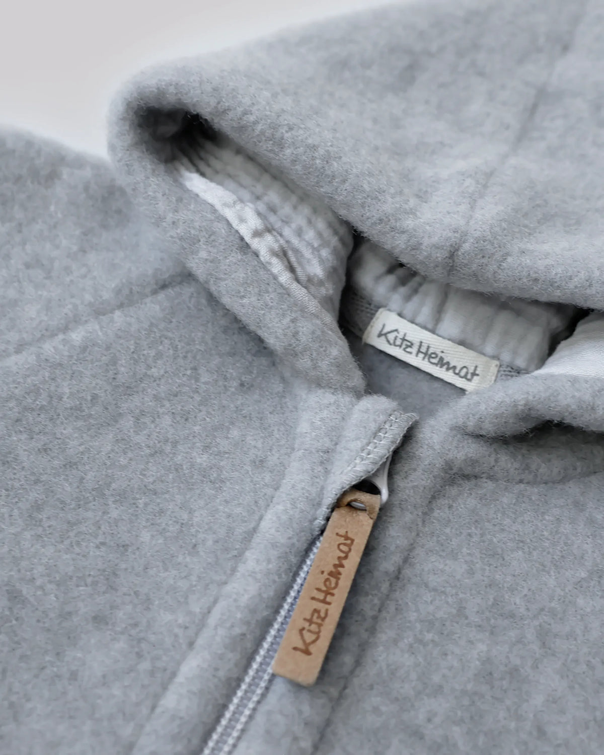 Nahaufnahme des Kitz Heimat Wollfleece-Overalls Sky Grey/Grey mit flauschiger Wollstruktur, hell gefütterter Kapuze und Reißverschluss mit Markenanhänger.
