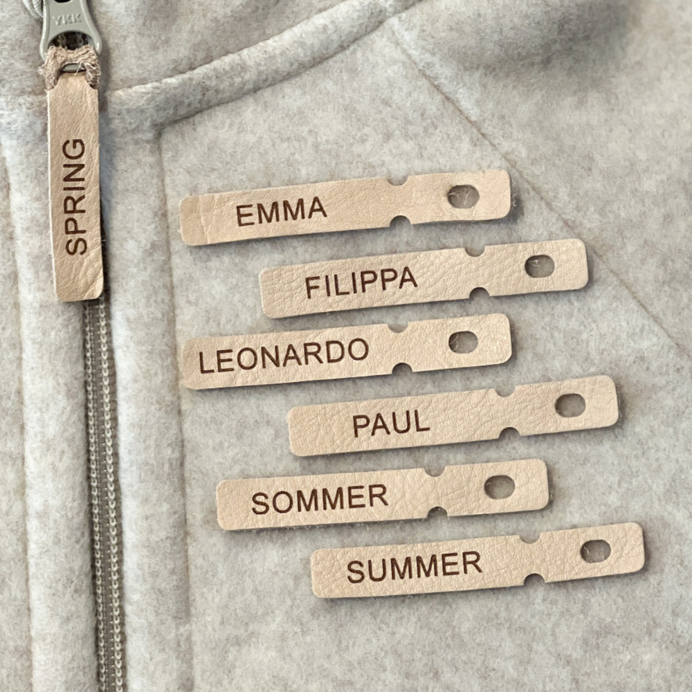 6 Lederanhänger mit verschiedenen Namen auf einem Nude farbenen Overall liegend.