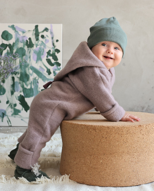 Baby trägt einen Merino Overall in Dusty Lilac und Mütze in Pine und stützt sich beim Stehen auf einem Hocker ab.